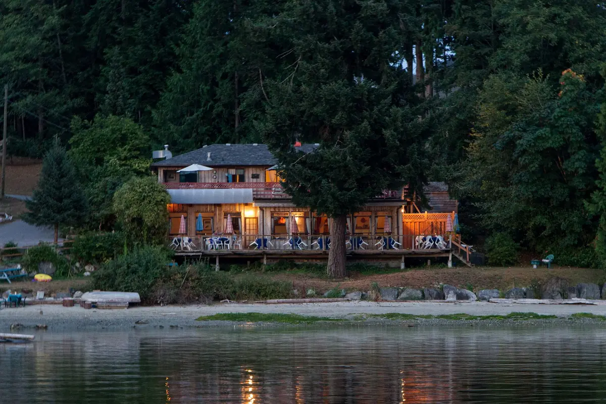 Beleuchtete Lodge am Wasser zwischen hohen Bäumen oberhalb eines Strandes in der Dämmerung